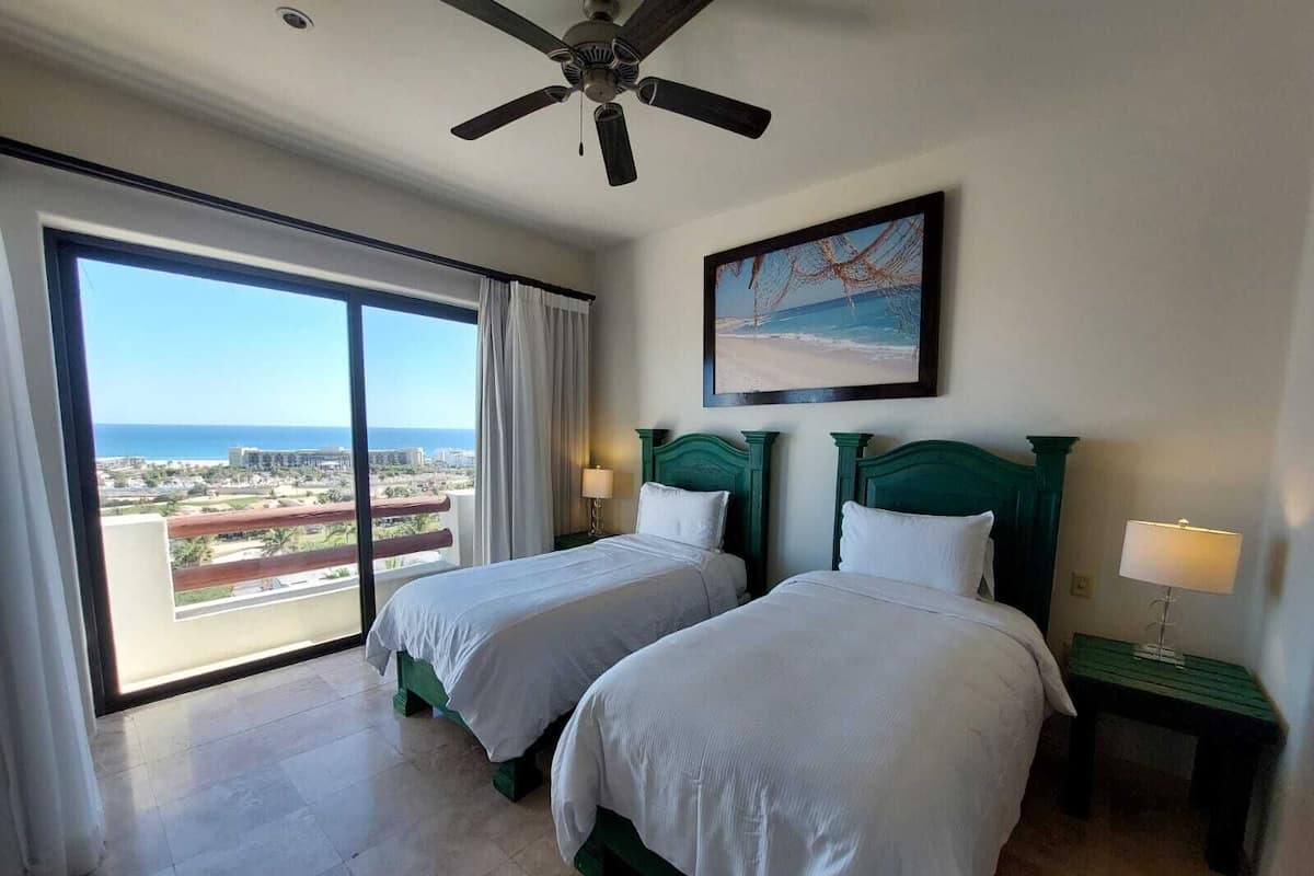 Une chambre d’hôtel avec deux lits, un ventilateur de plafond, un tableau représentant une plage et une vue sur l’océan et les bâtiments.
