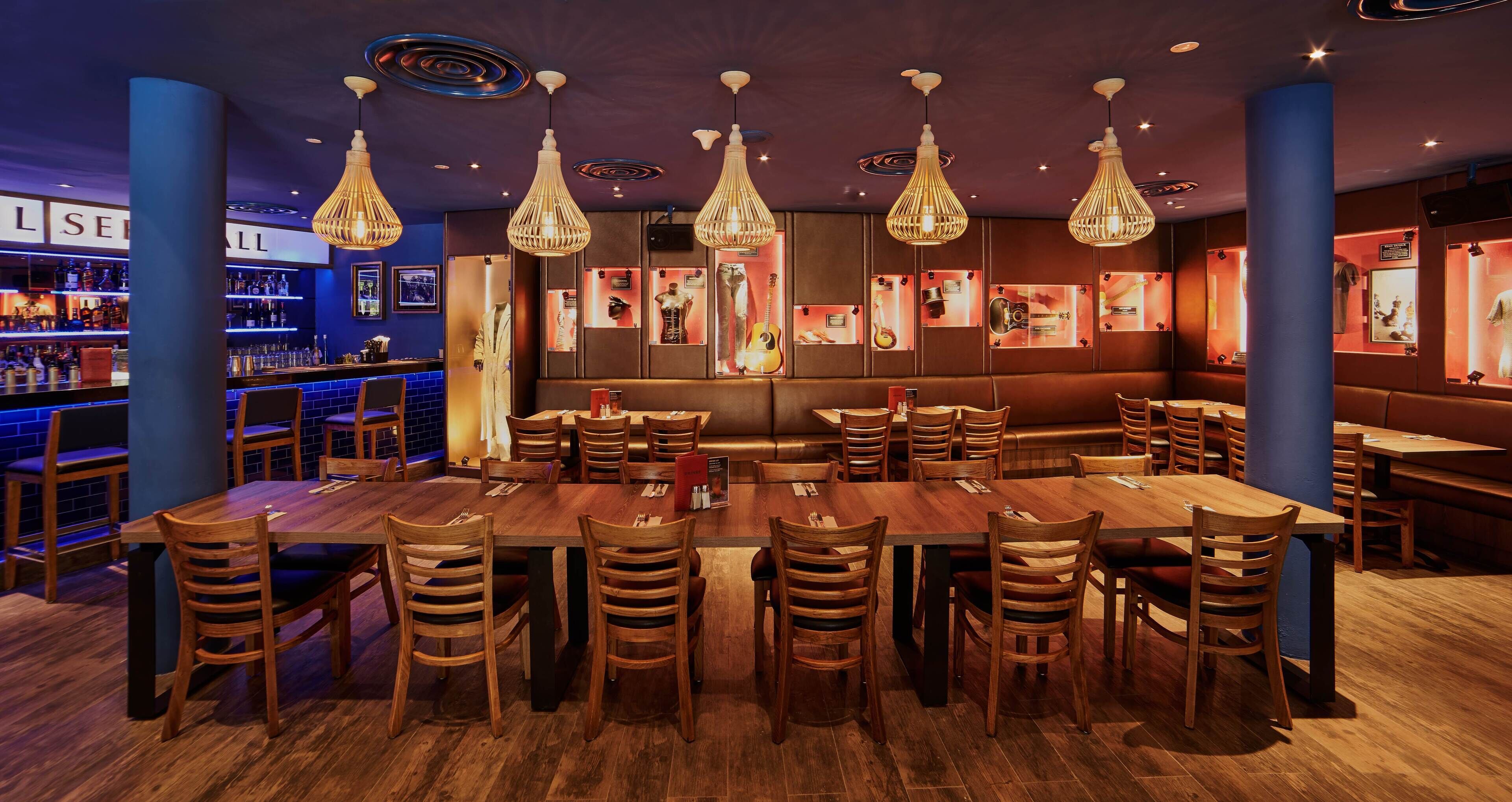 A restaurant with a long wooden table surrounded by chairs, hanging pendant lights, and framed pictures on the walls.