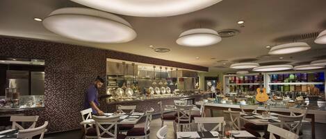 A modern restaurant with a central bar area, white chairs, and a variety of dishes displayed.