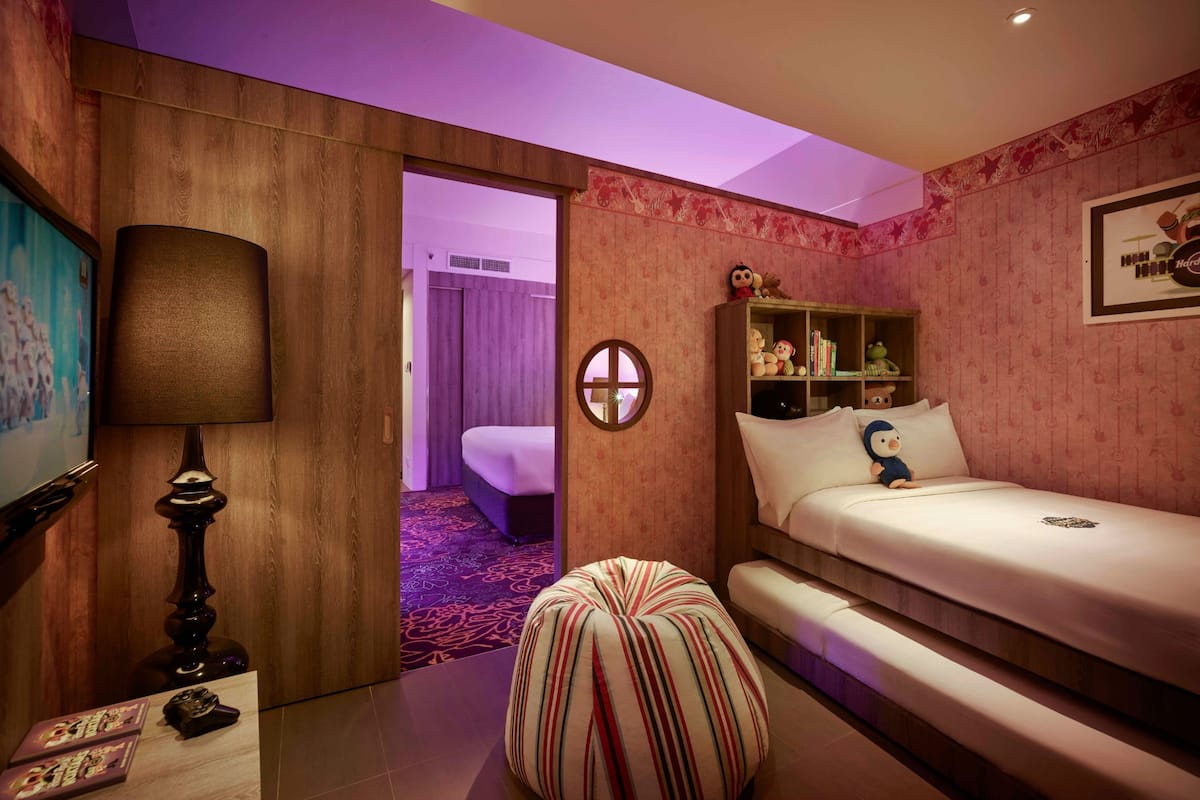 A hotel room with a bed, a small table with a lamp, a round ottoman, a shelf with decorative items, and a painting on the wall.