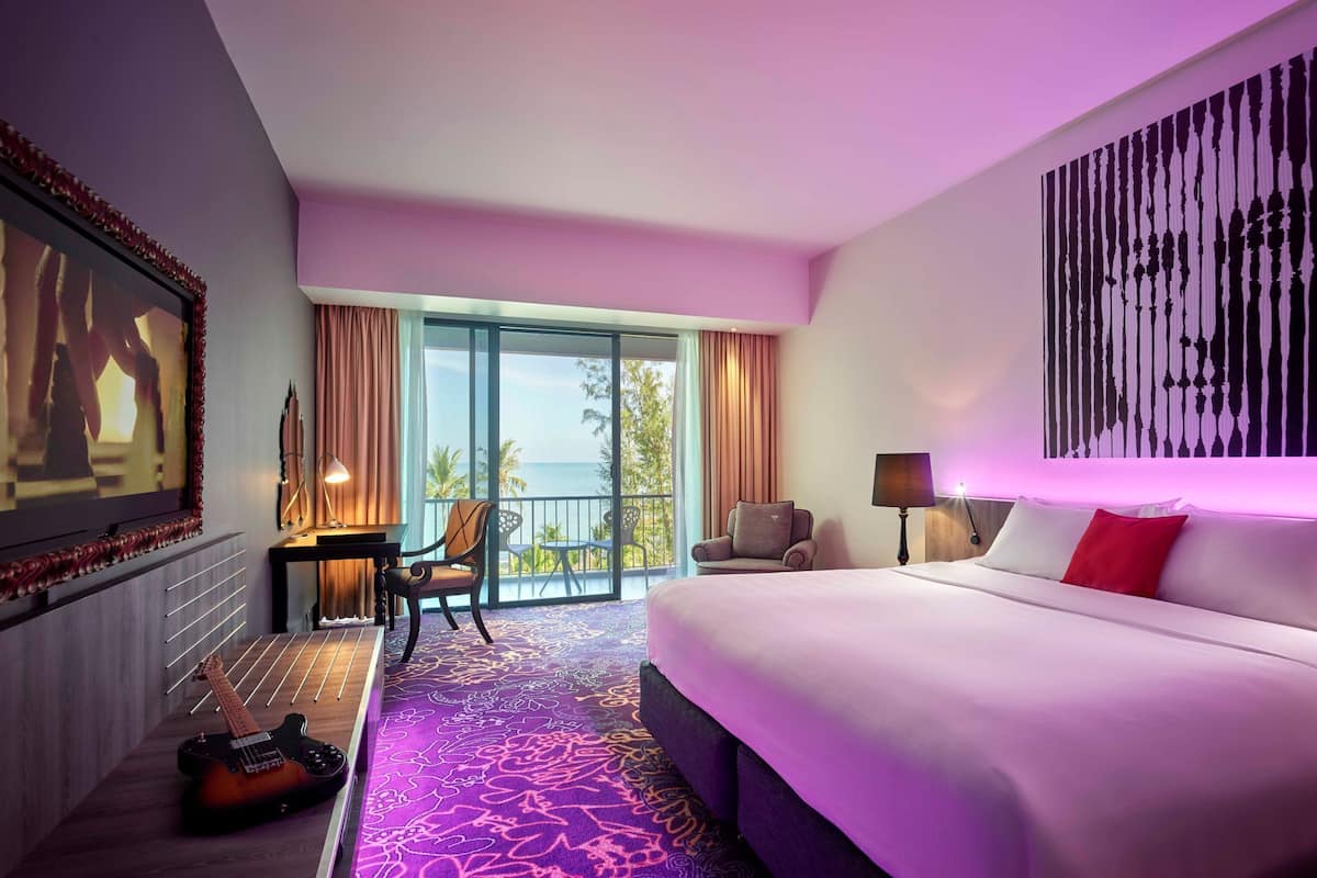 A hotel room with a large bed, a desk with a chair, a guitar, a balcony with a view of the ocean, and a large painting on the wall.