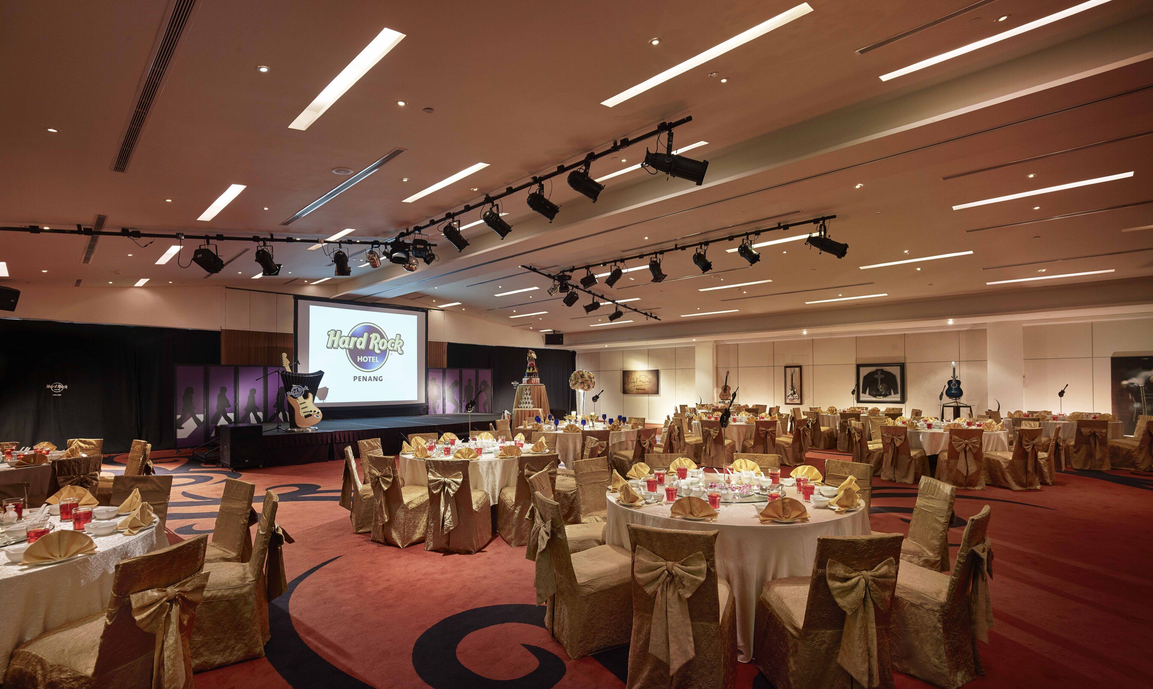 Una sala de banquetes amb taules rodones, cadires amb llaços i un escenari amb una pantalla de projector que mostra "Hard Rock Hotel Penang".
