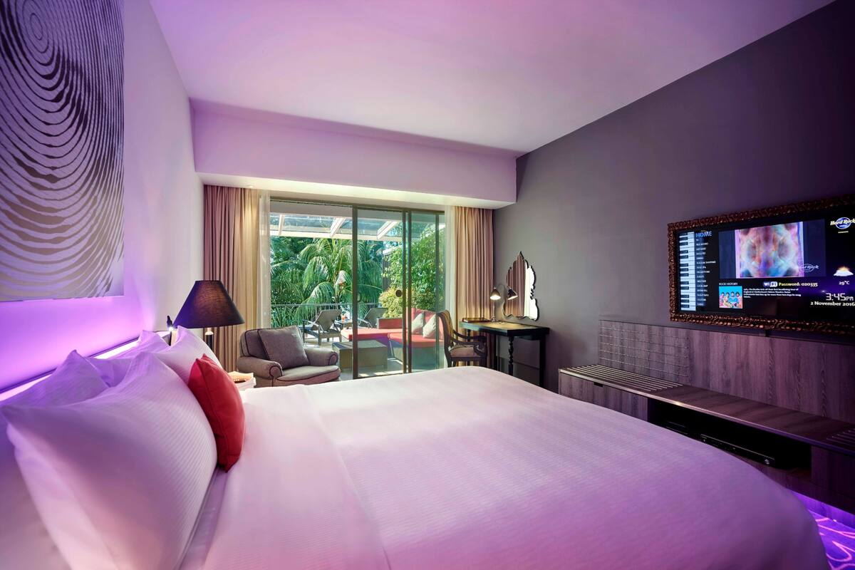 A modern hotel room with a large bed, a TV mounted on the wall, and a balcony with outdoor seating.