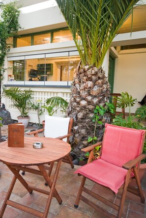 A patio with a round wooden table and two chairs, one with a red cushion. A tall palm tree is in the background.