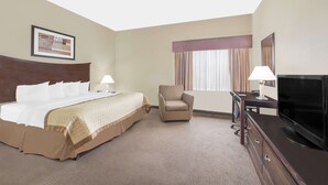 A hotel room with a large bed, a chair, a desk with a TV, and a window with curtains. - Baymont by Wyndham Snyder (Snyder)