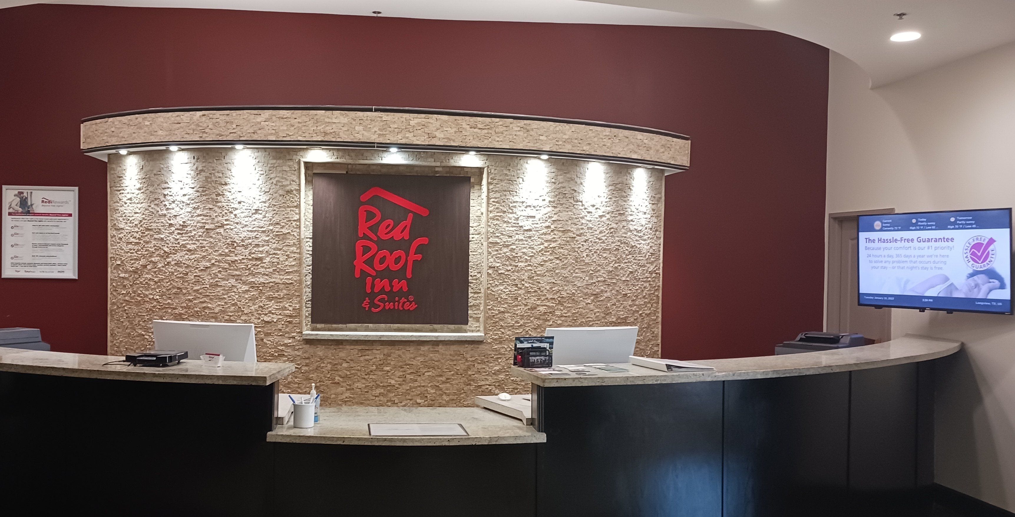 Mostrador de recepción en Red Roof Inn Suites, con una pared de textura pétrea, una pantalla digital que muestra información y un cartel enmarcado de Red Rewards.