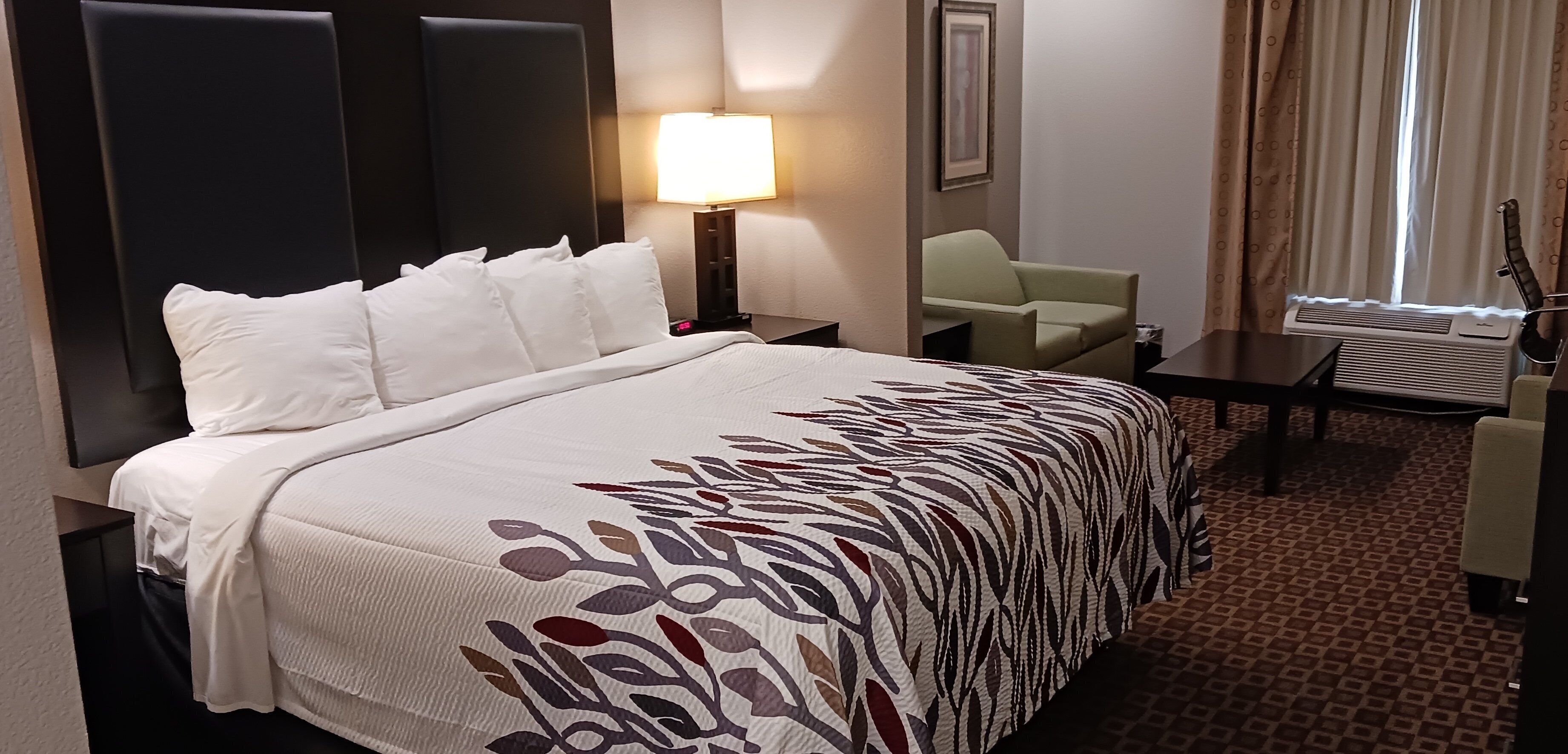 A hotel room with a bed, a desk, a chair, a lamp, and an air conditioning unit.