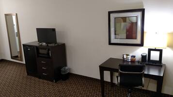 Una habitación de hotel con escritorio, silla, televisor sobre una cómoda, un cuadro en la pared y un cubo de basura.