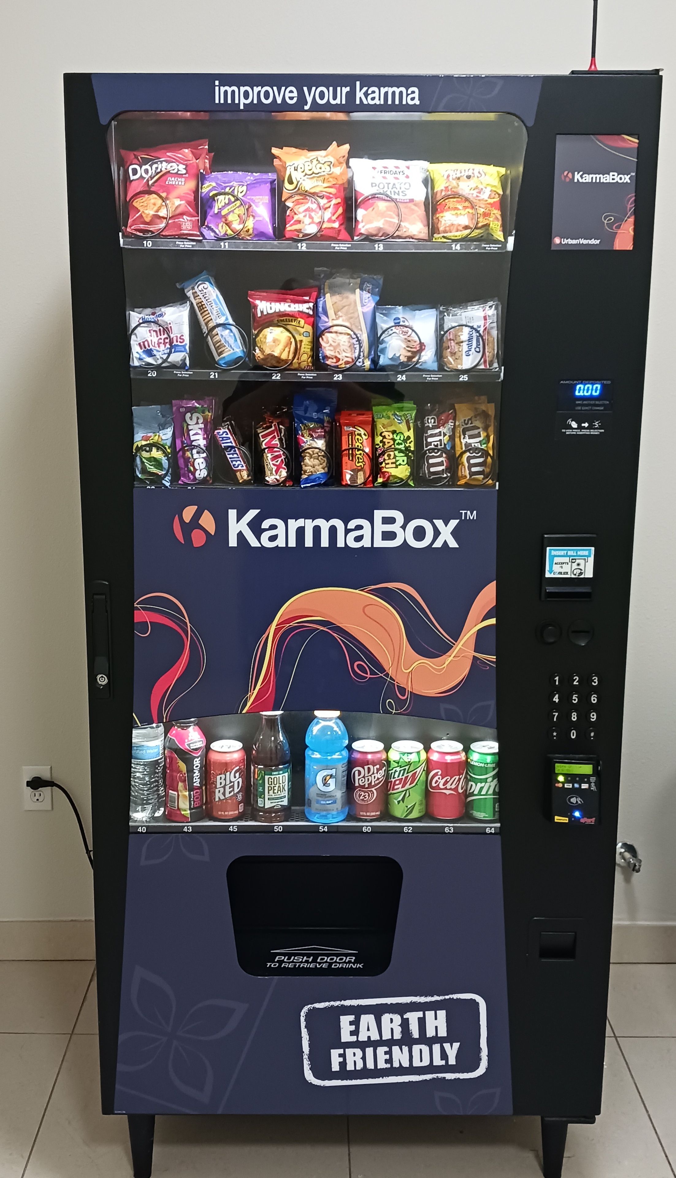 Máquina expendedora de la marca 'KarmaBox' con el lema 'mejora tu karma'. Tiene una pantalla digital y un teclado para seleccionar.
