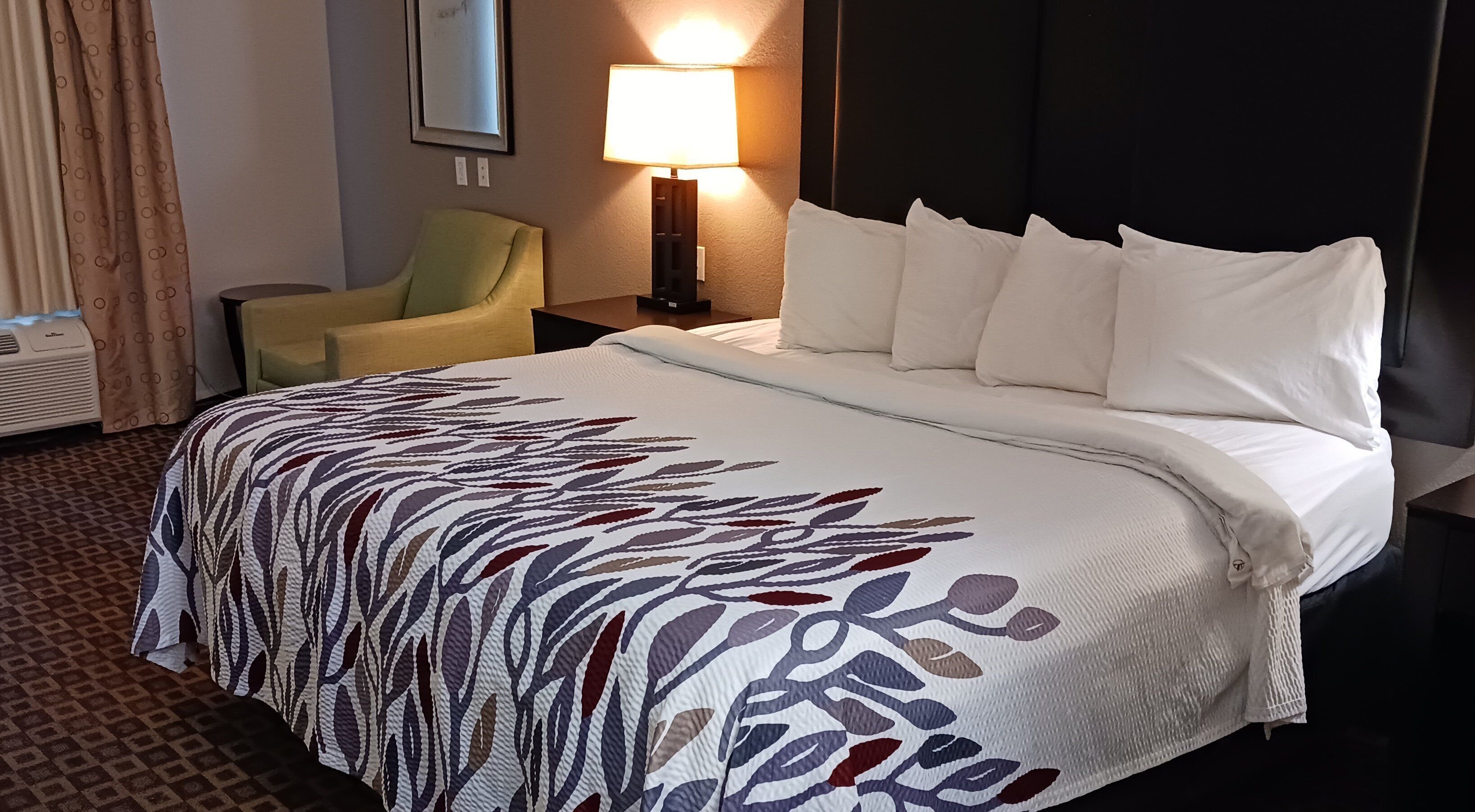 A hotel room with a bed, a chair, a nightstand with a lamp, and a wall-mounted picture.