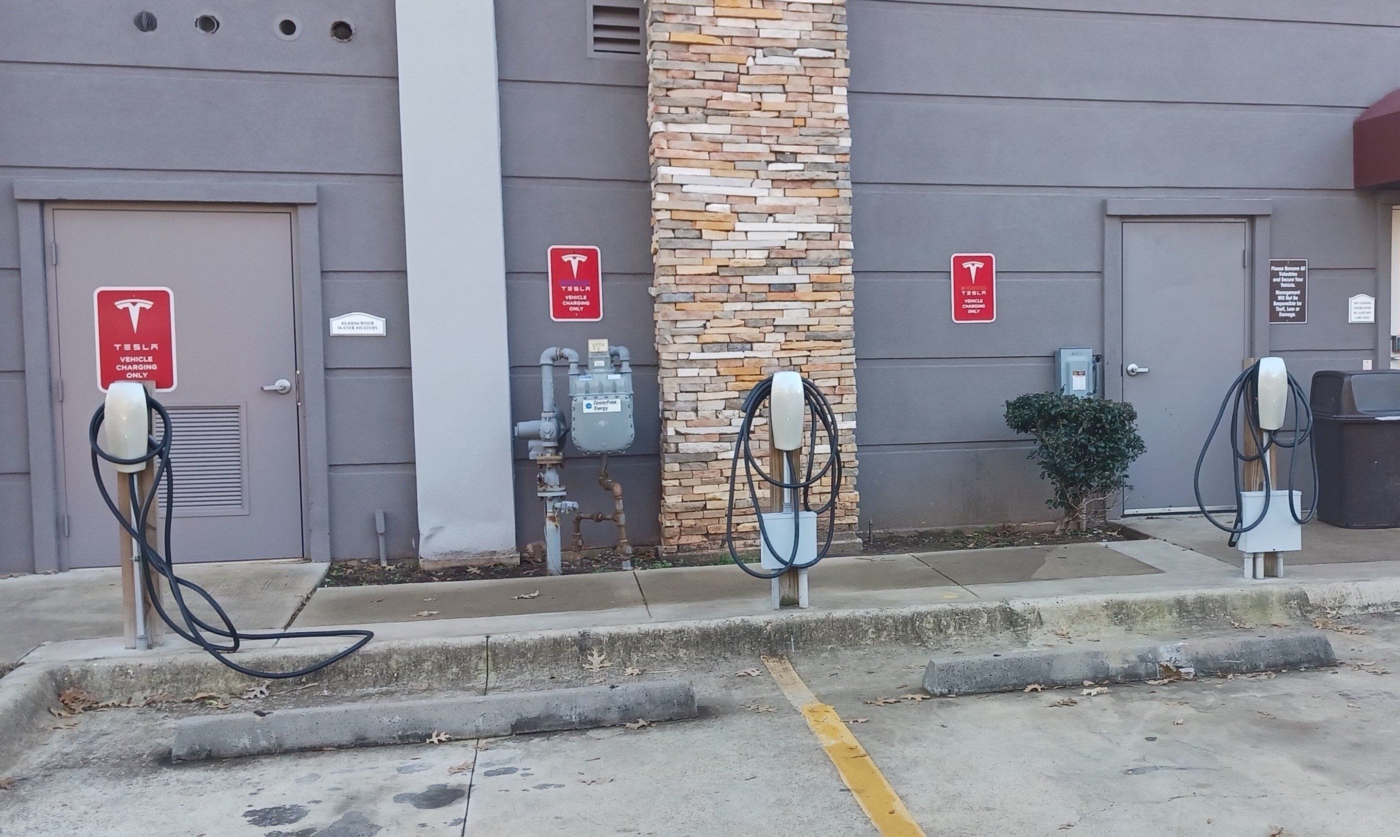 A parking area with three Tesla charging stations, a water heater, and a building with a stone facade.