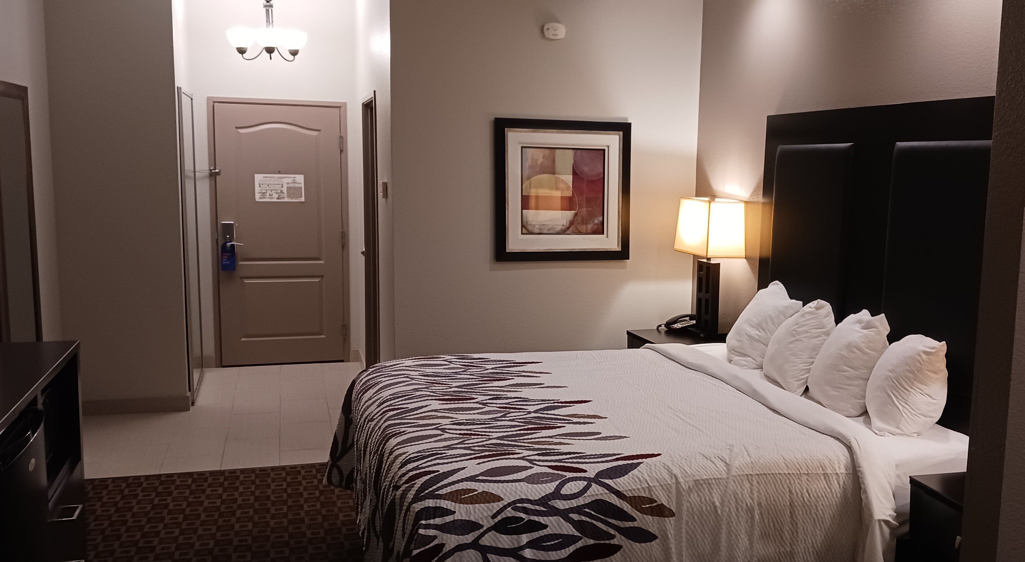 Een hotelkamer met een bed, een nachtkastje met een lamp, een deur en een ingelijst schilderij aan de muur.