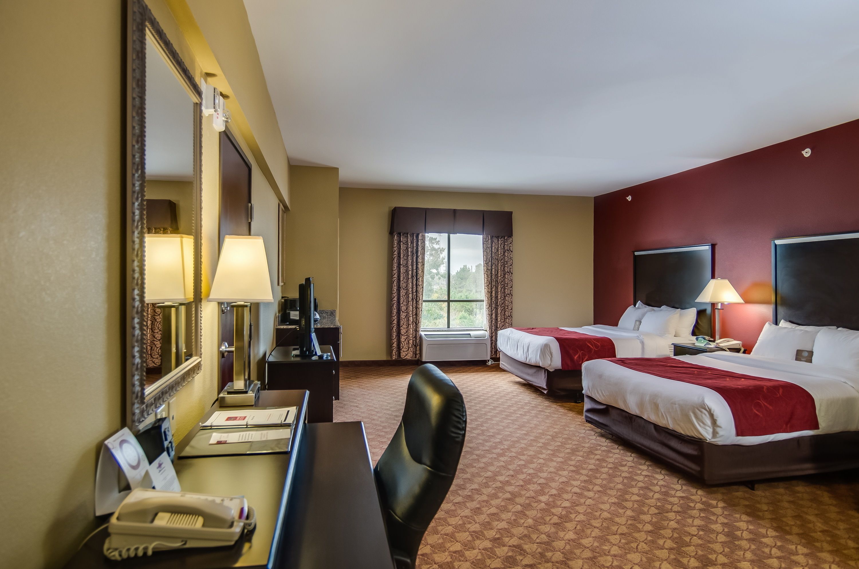 Une chambre d’hôtel avec deux lits, un bureau avec un téléphone, un miroir et une fenêtre avec des rideaux.