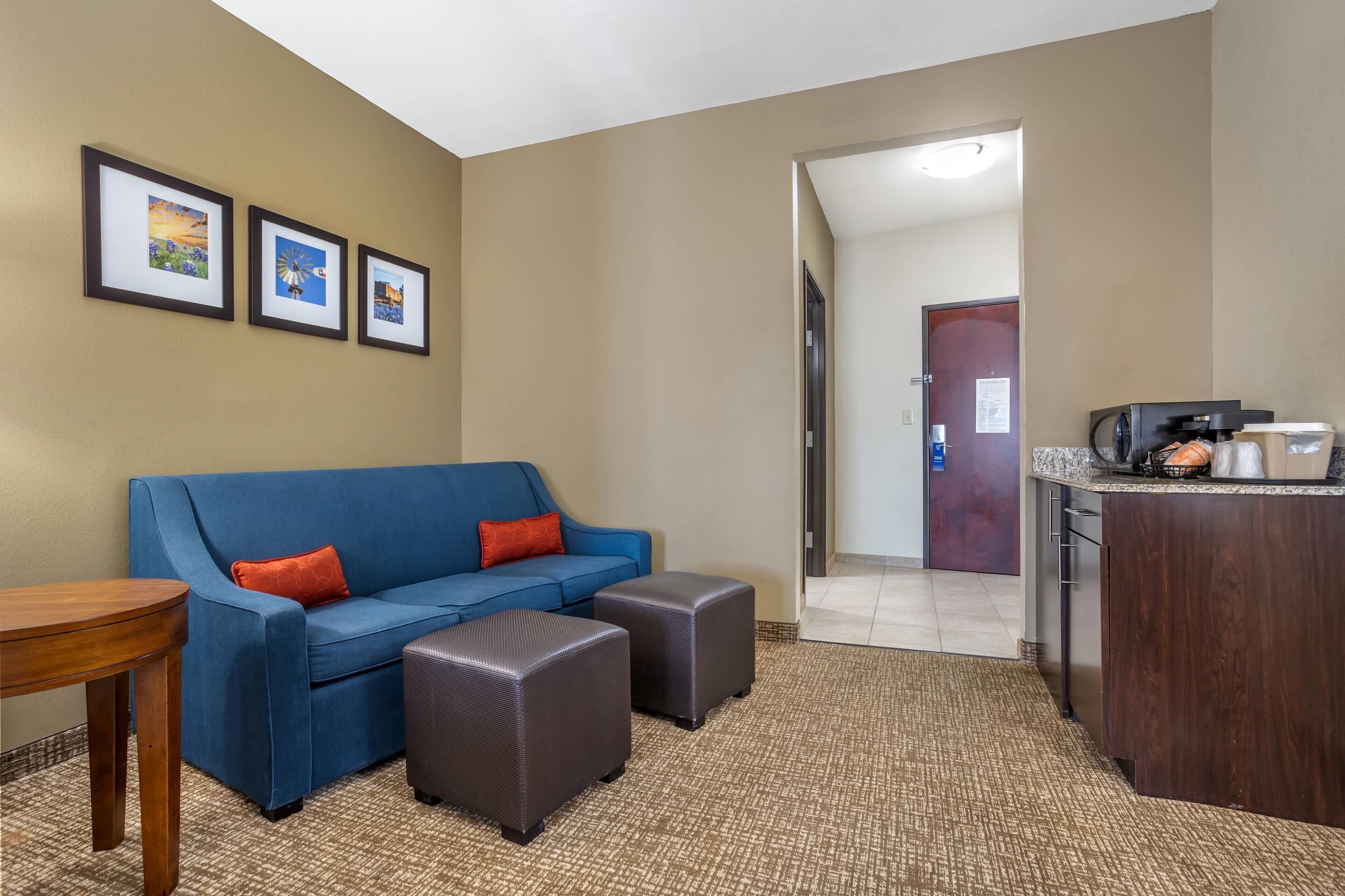 A hotel room with a blue sofa, two ottomans, a small wooden table, a kitchenette with a coffee maker, and framed pictures on the wall.