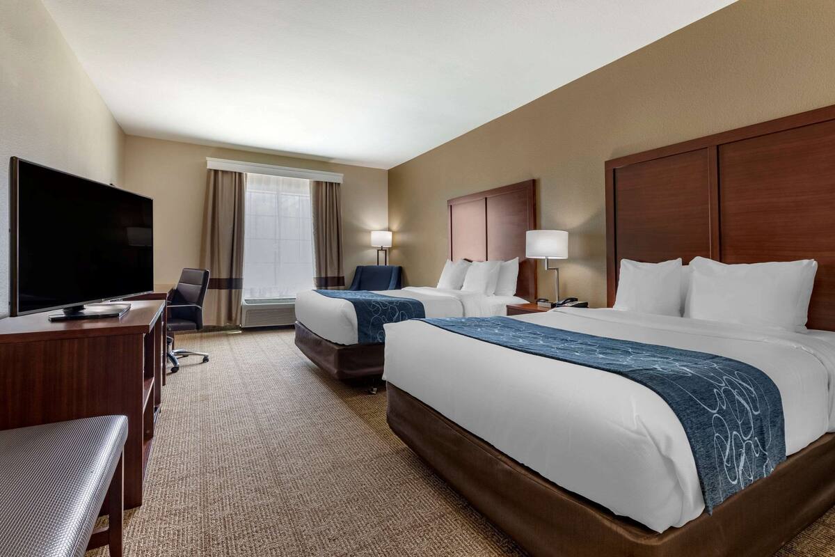 A hotel room with two beds, a desk with a TV, a chair, a lamp, and a large window with curtains.