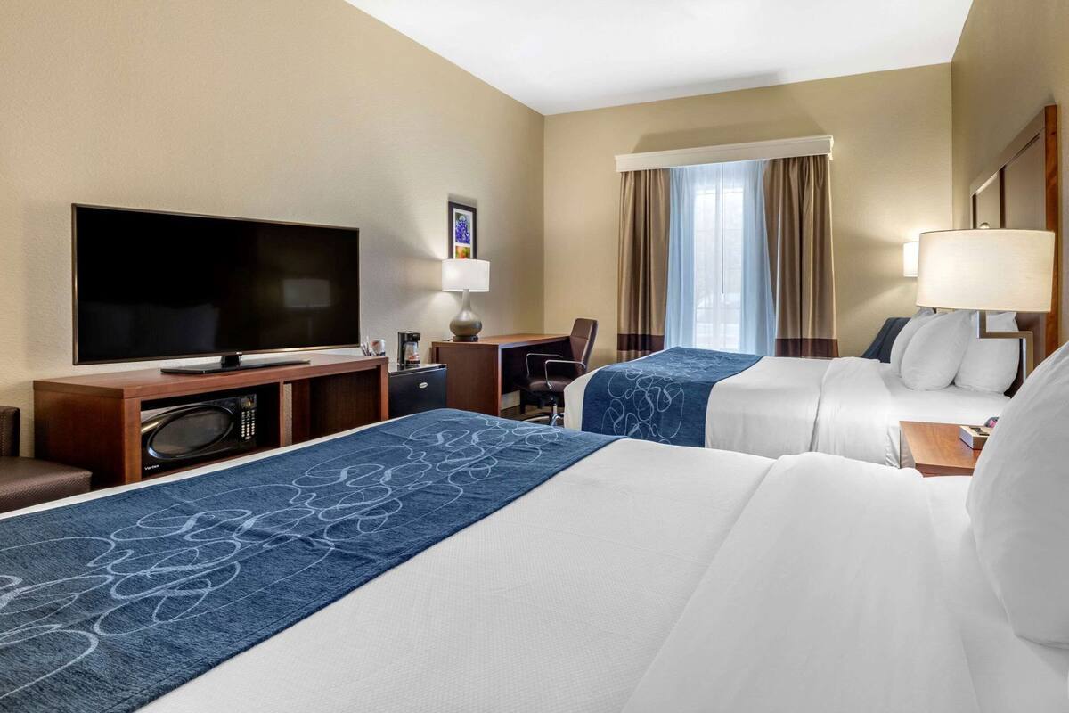 A hotel room with two beds, a TV, a desk with a computer, and a window with curtains.