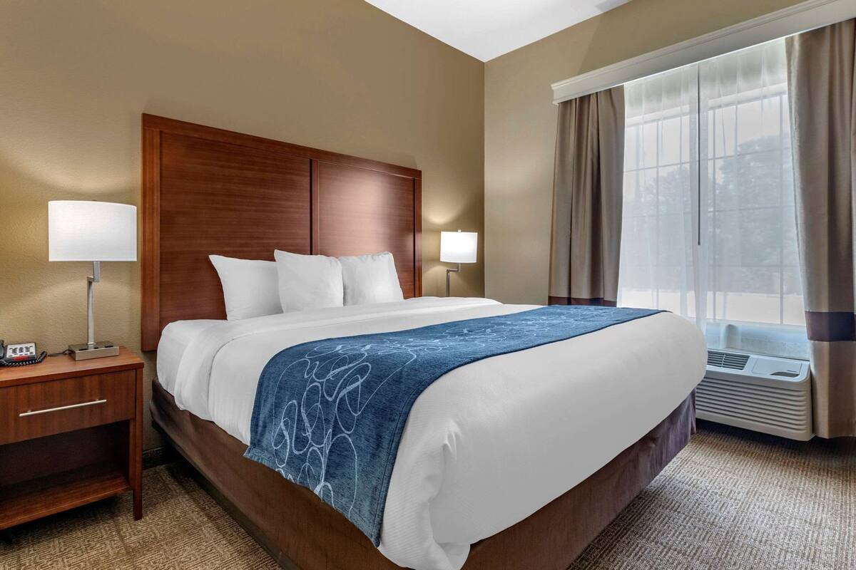 A hotel room with a large bed, two bedside lamps, a wooden headboard, a window with curtains, and a carpeted floor.