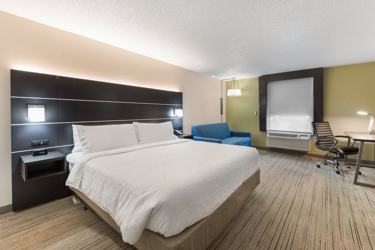 A hotel room with a large bed, a desk, a chair, and a window with blinds.