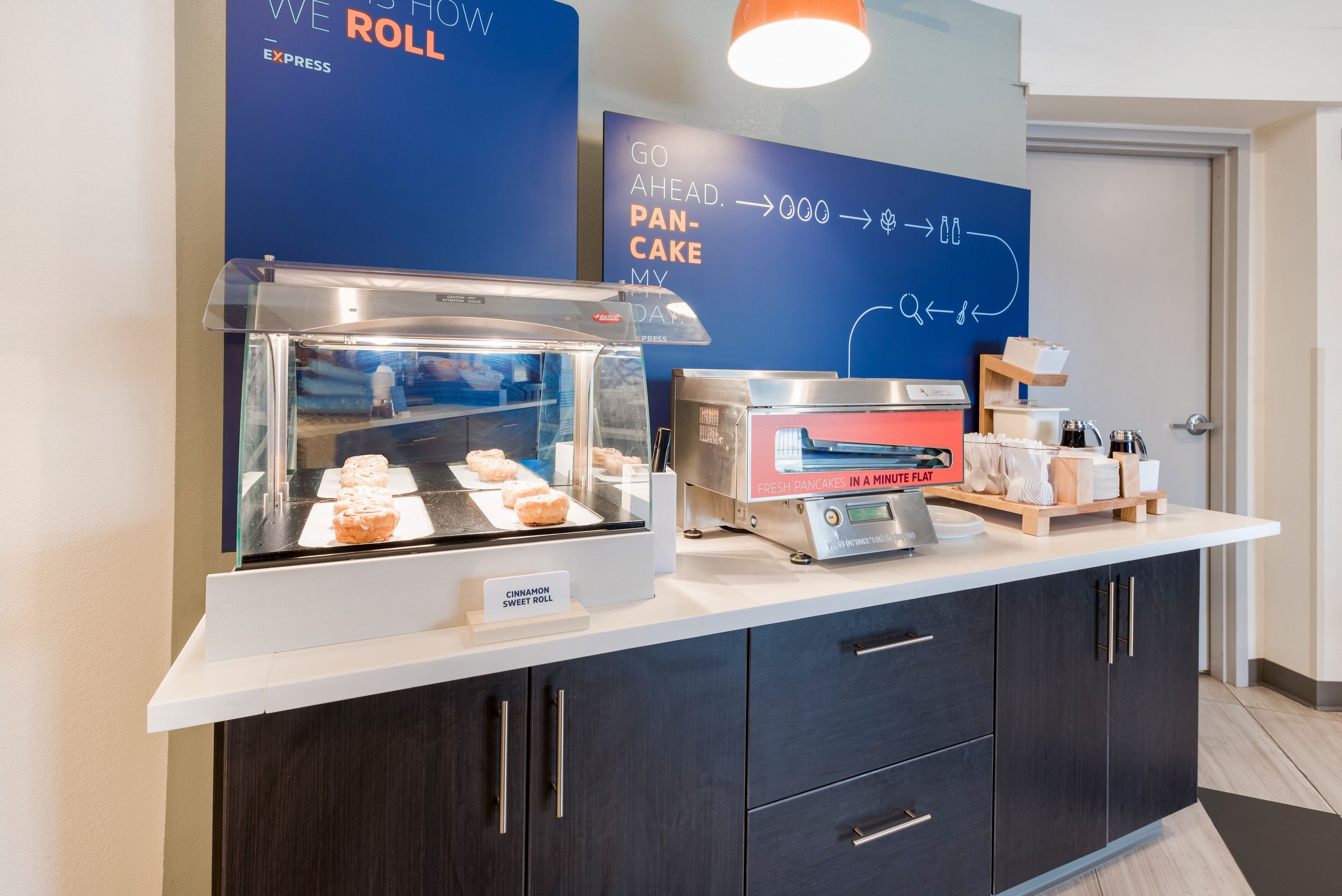 Un coin petit-déjeuner avec un présentoir de viennoiseries, une machine à panini et un panneau indiquant que des crêpes fraîches sont disponibles.