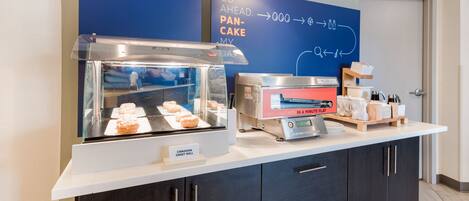 Un coin petit-déjeuner avec un présentoir de viennoiseries, une machine à panini et un panneau indiquant que des crêpes fraîches sont disponibles.