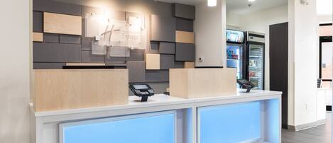 A hotel reception desk with a modern design, a digital payment terminal, and a refrigerator stocked with beverages.