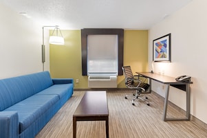 Ett hotellrum med en blå soffa, ett skrivbord med en telefon, en stol, en lampa och ett fönster med persienner.
