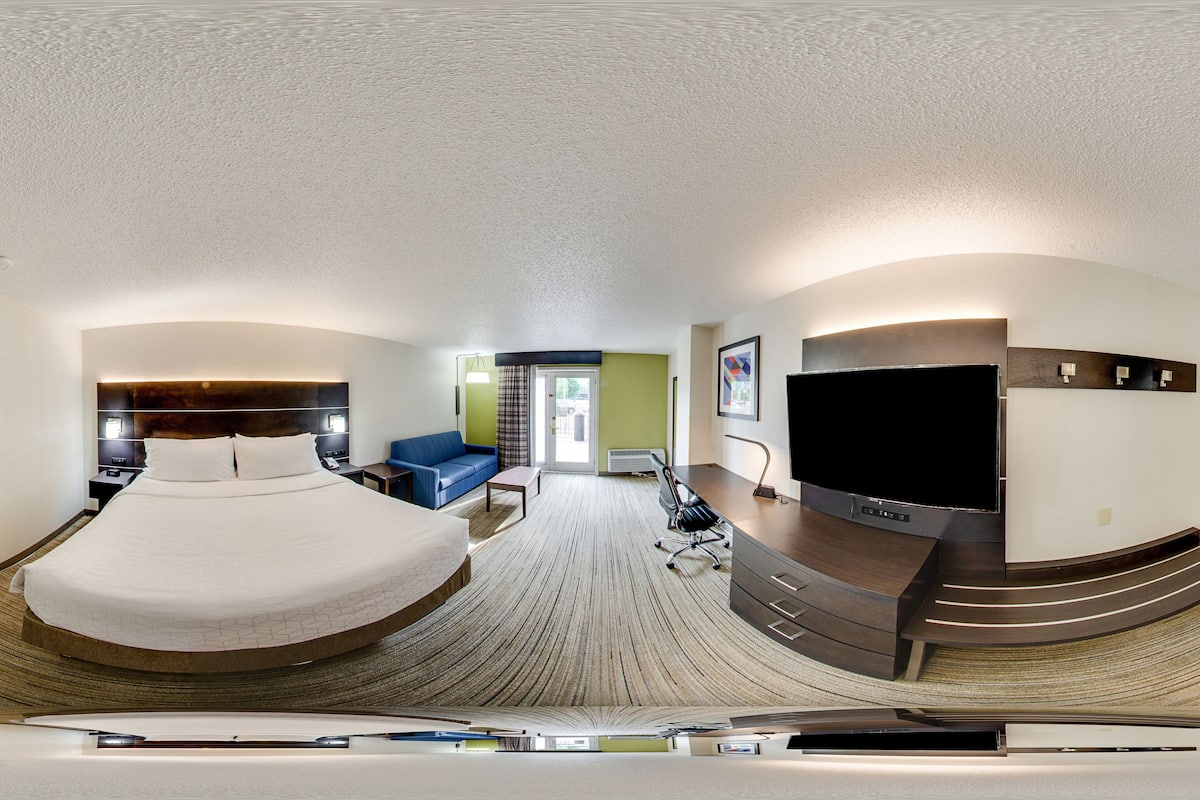 Vue panoramique d’une chambre d’hôtel comprenant un grand lit, un bureau, une télévision et un coin salon avec un canapé et des fauteuils.