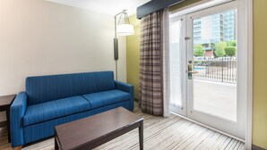 A room with a blue sofa, a small wooden table, a floor lamp, and a sliding glass door leading to an outdoor area with a view of buildings and trees. - Holiday Inn Express Columbus Downtown by IHG (Columbus)