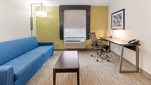 A hotel room with a blue sofa, a wooden table, a desk with a chair, a standing lamp, an air conditioning unit, and a framed picture on the wall. - Holiday Inn Express Columbus Downtown by IHG (Columbus)