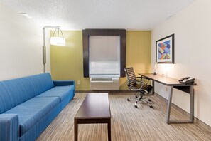A hotel room with a blue sofa, a wooden table, a desk with a chair, a standing lamp, an air conditioning unit, and a framed picture on the wall. - Holiday Inn Express Columbus Downtown by IHG (Columbus)