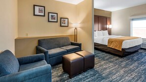 Une chambre d’hôtel avec un canapé, un pouf et un lit recouvert d’une couverture marron. Des tableaux encadrés ornent le mur, et une lampe sur pied est posée au sol.