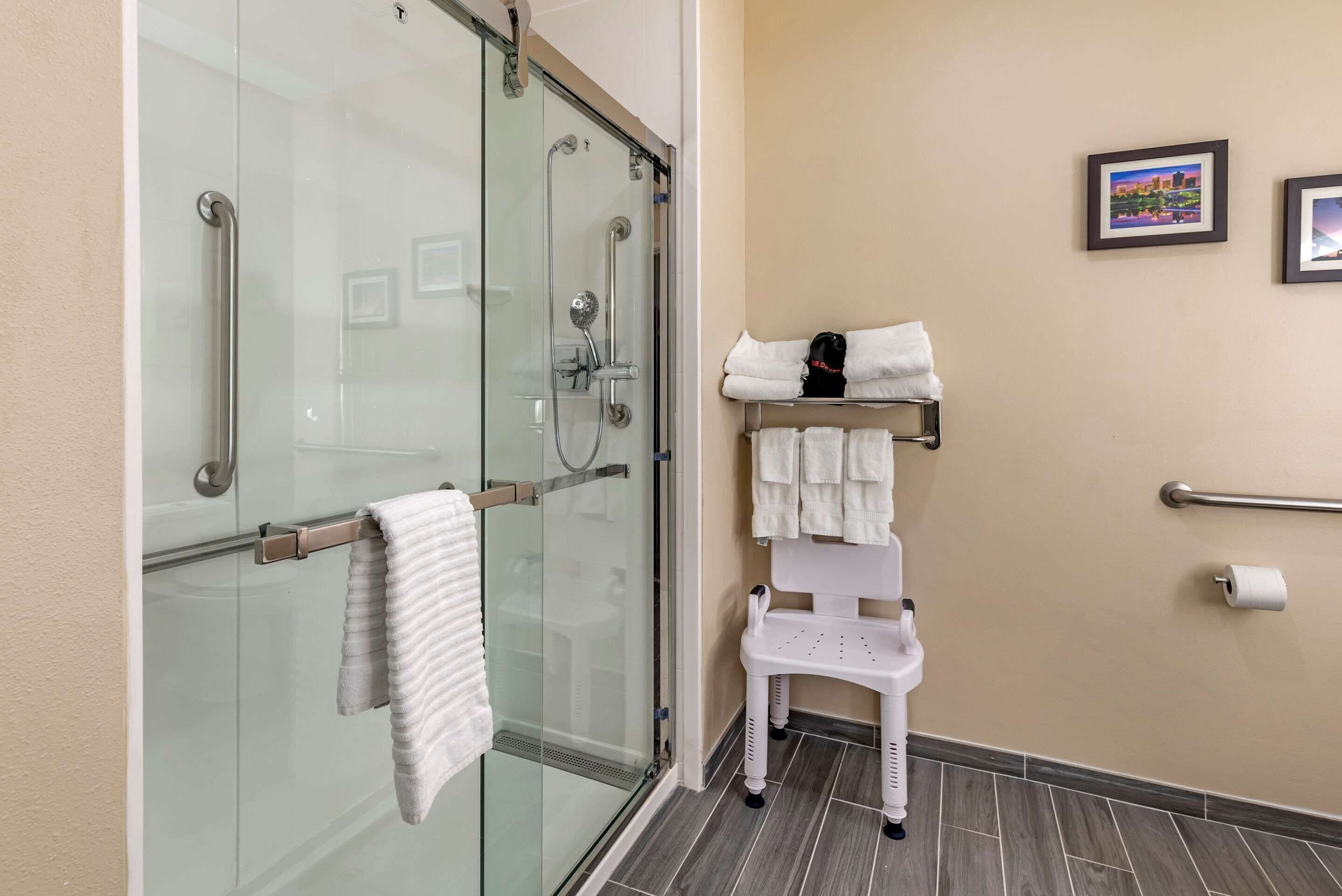 Une salle de bain dotée d’une douche à parois de verre, d’un porte-serviettes blanc, d’une barre d’appui blanche et d’une étagère contenant des serviettes et une casquette noire.