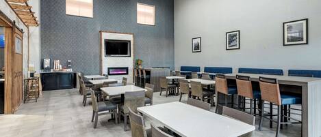 A modern dining area with multiple tables and chairs, a kitchen area with a counter, and a large TV screen.