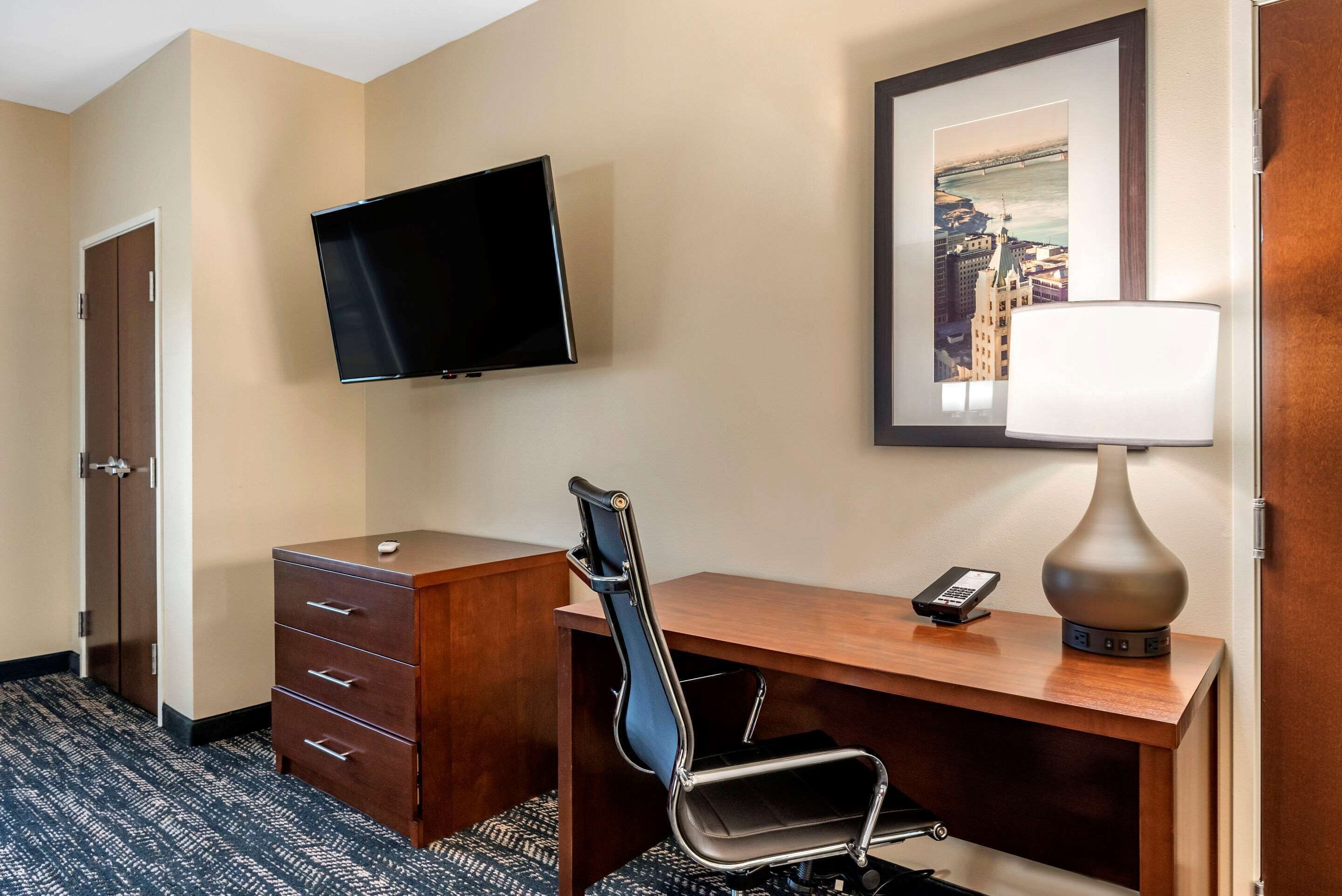 Habitación de hotel con escritorio, silla, televisor, lámpara y una fotografía enmarcada de un paisaje urbano.