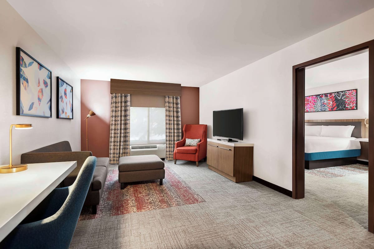 Une chambre d’hôtel avec un lit, un bureau et une chaise, une télévision, un fauteuil rouge et une grande fenêtre avec des rideaux.