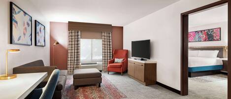 A hotel room with a bed, a desk with a chair, a television, a red armchair, and a large window with curtains.