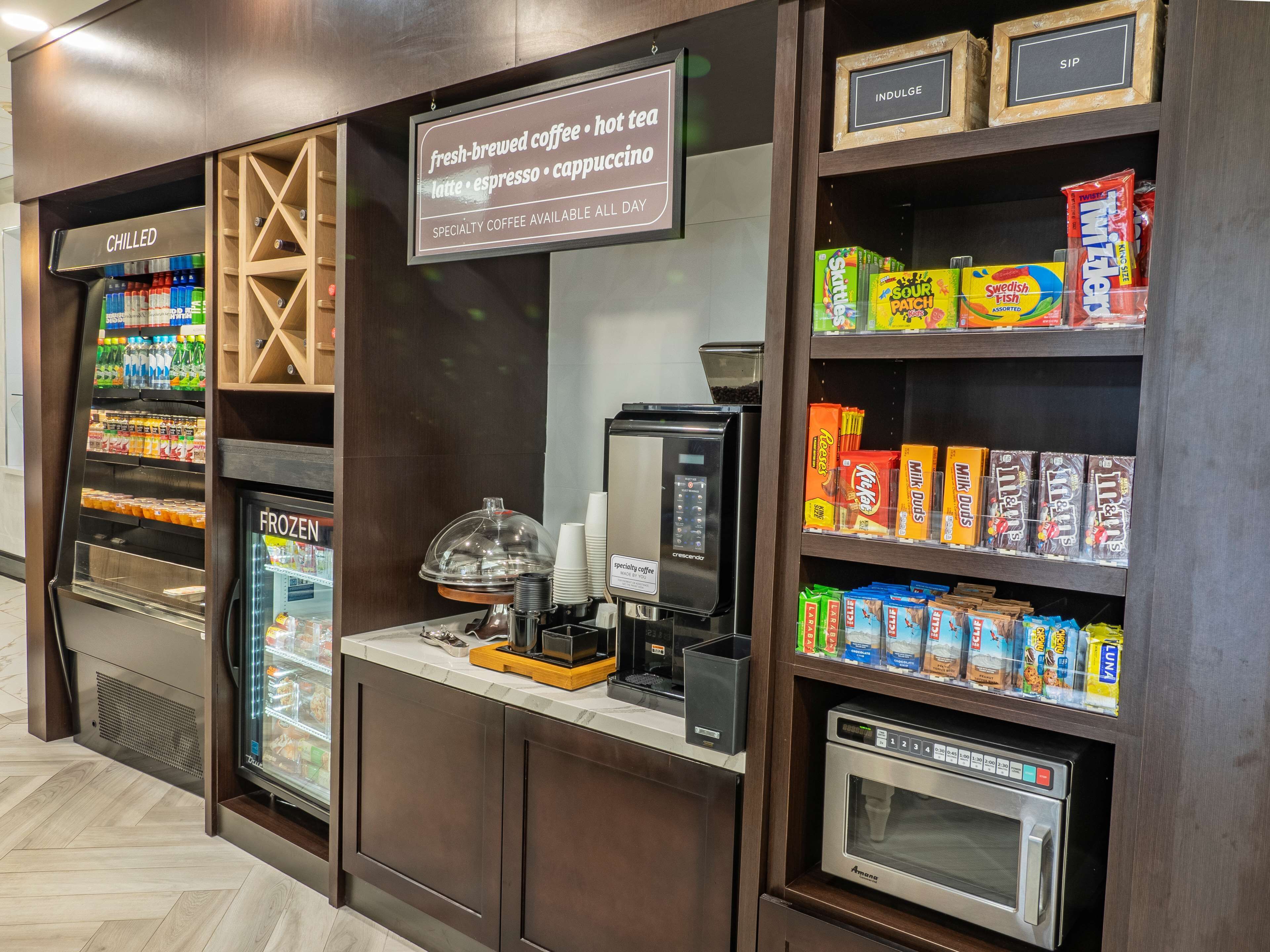 Una estación de café con bebidas frías y congeladas, una máquina de café, y una variedad de bocadillos y cartones de leche.