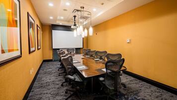 A conference room with a long table, chairs, a projector screen, and framed artwork on the walls.