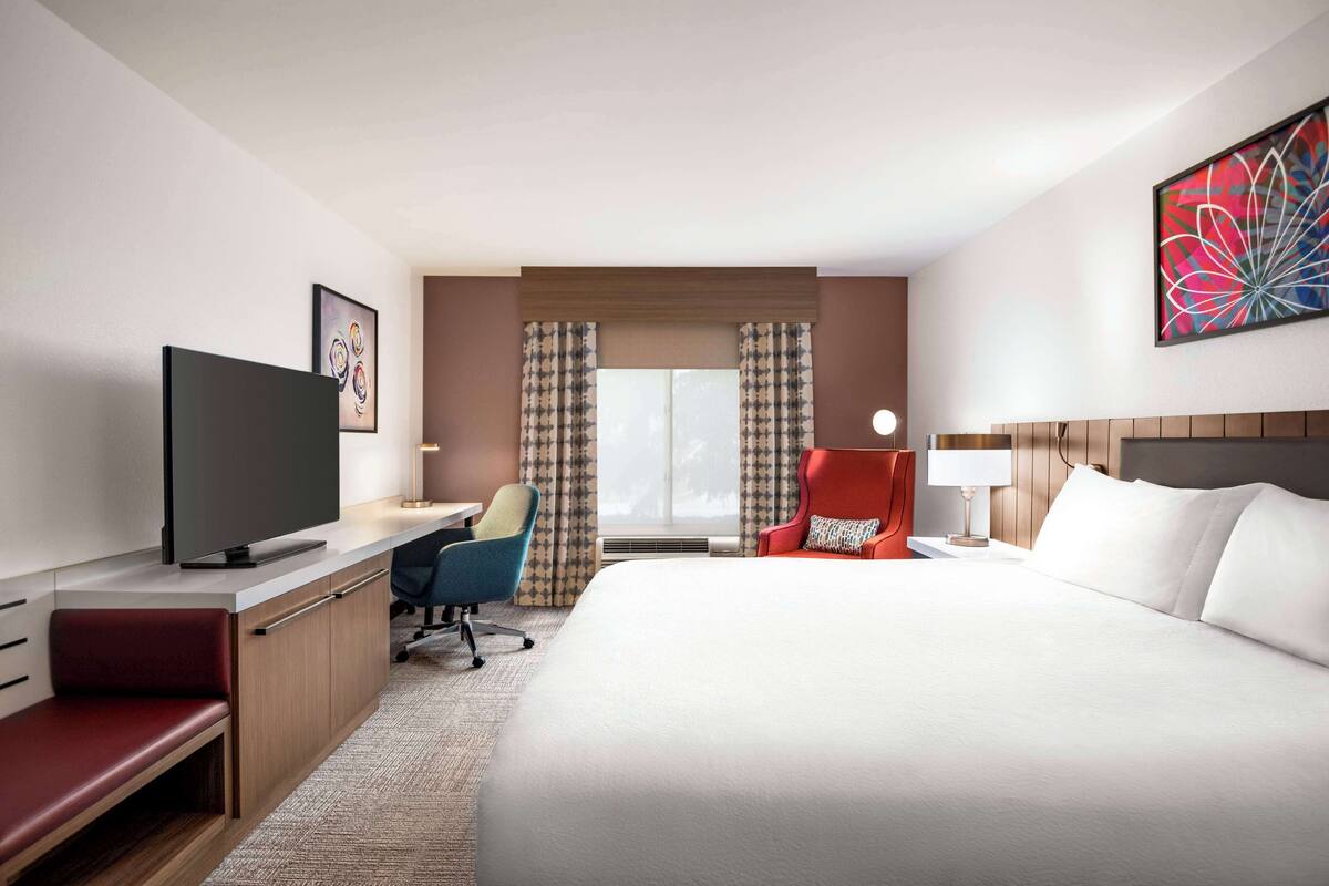 Une chambre d’hôtel avec un grand lit, un bureau avec une chaise, une télévision et une fenêtre avec des rideaux.