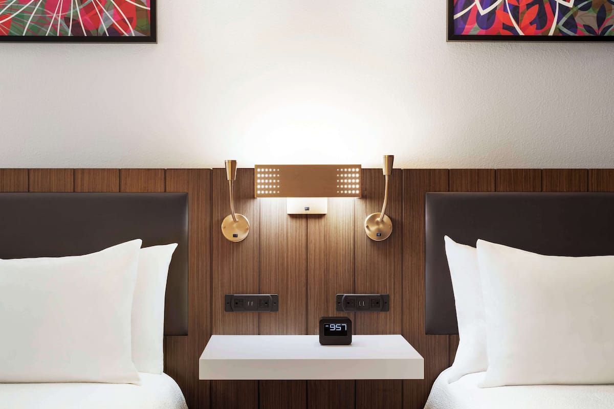 Une chambre d’hôtel avec deux lits, une table de chevet, une horloge numérique et des lampes fixées au mur.
