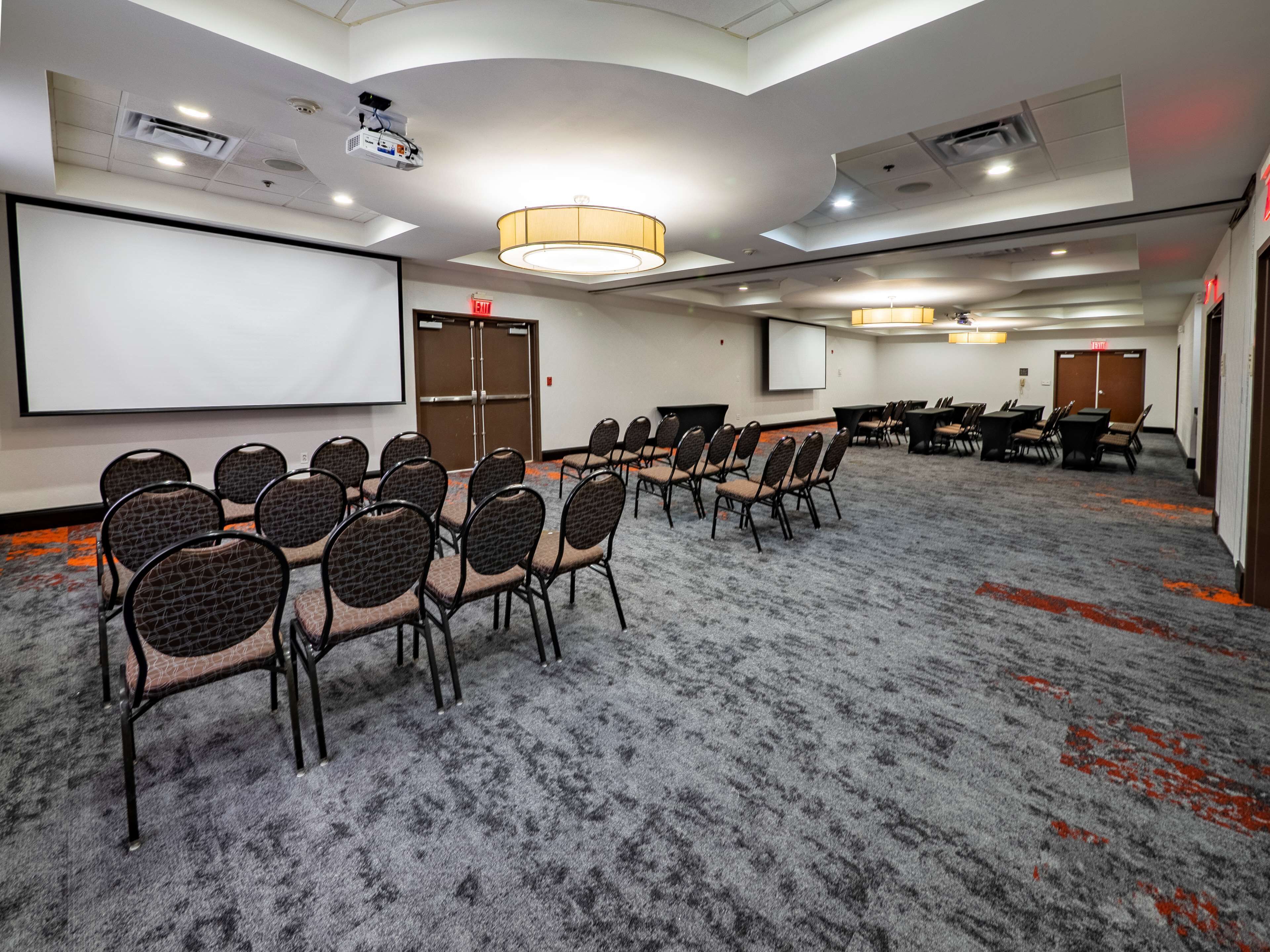 Un salón de conferencias con una pantalla grande, varias mesas y sillas dispuestas para reuniones.