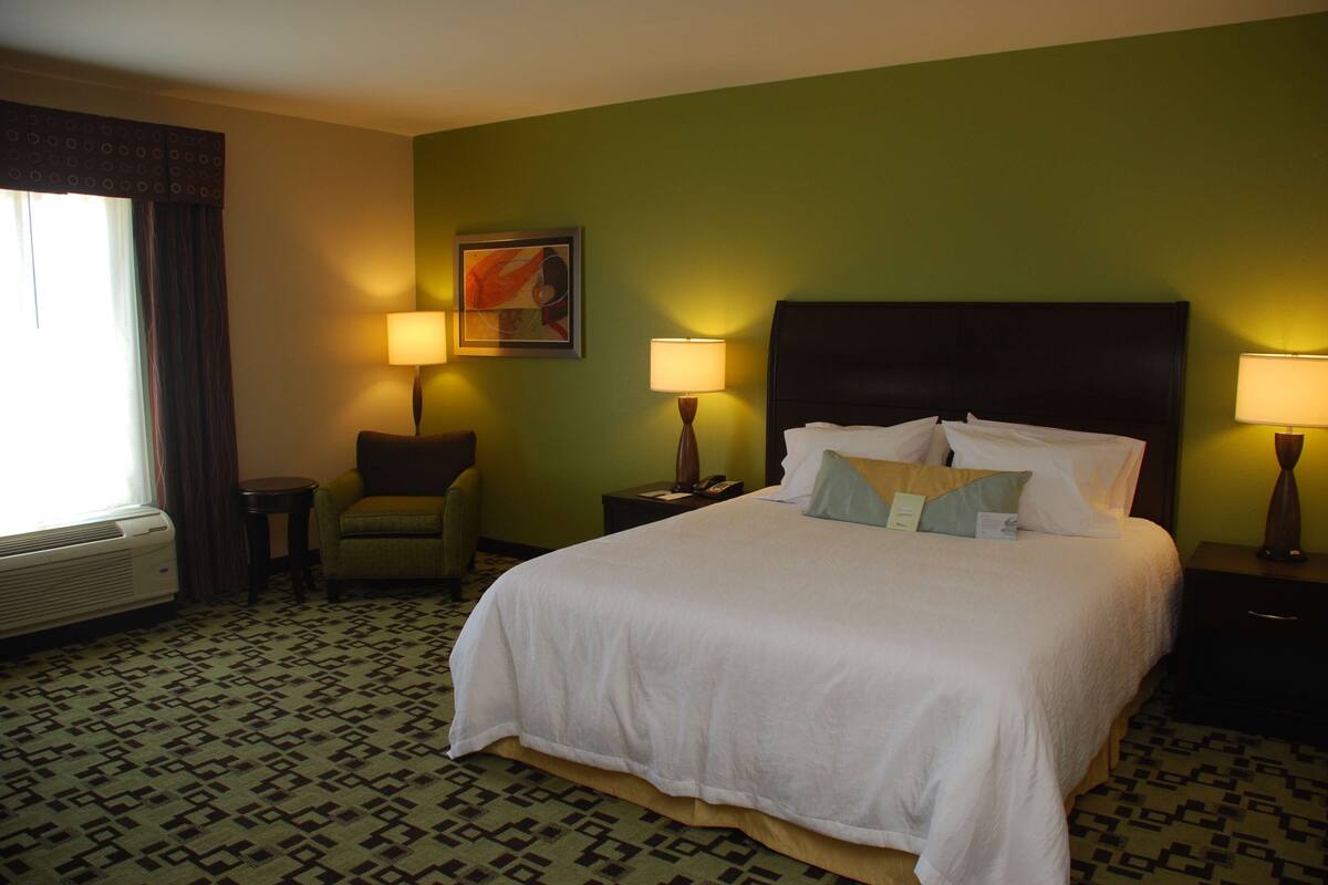 Habitación de hotel con una cama grande, dos mesitas de noche con lámparas, una silla y un ventanal con cortinas.