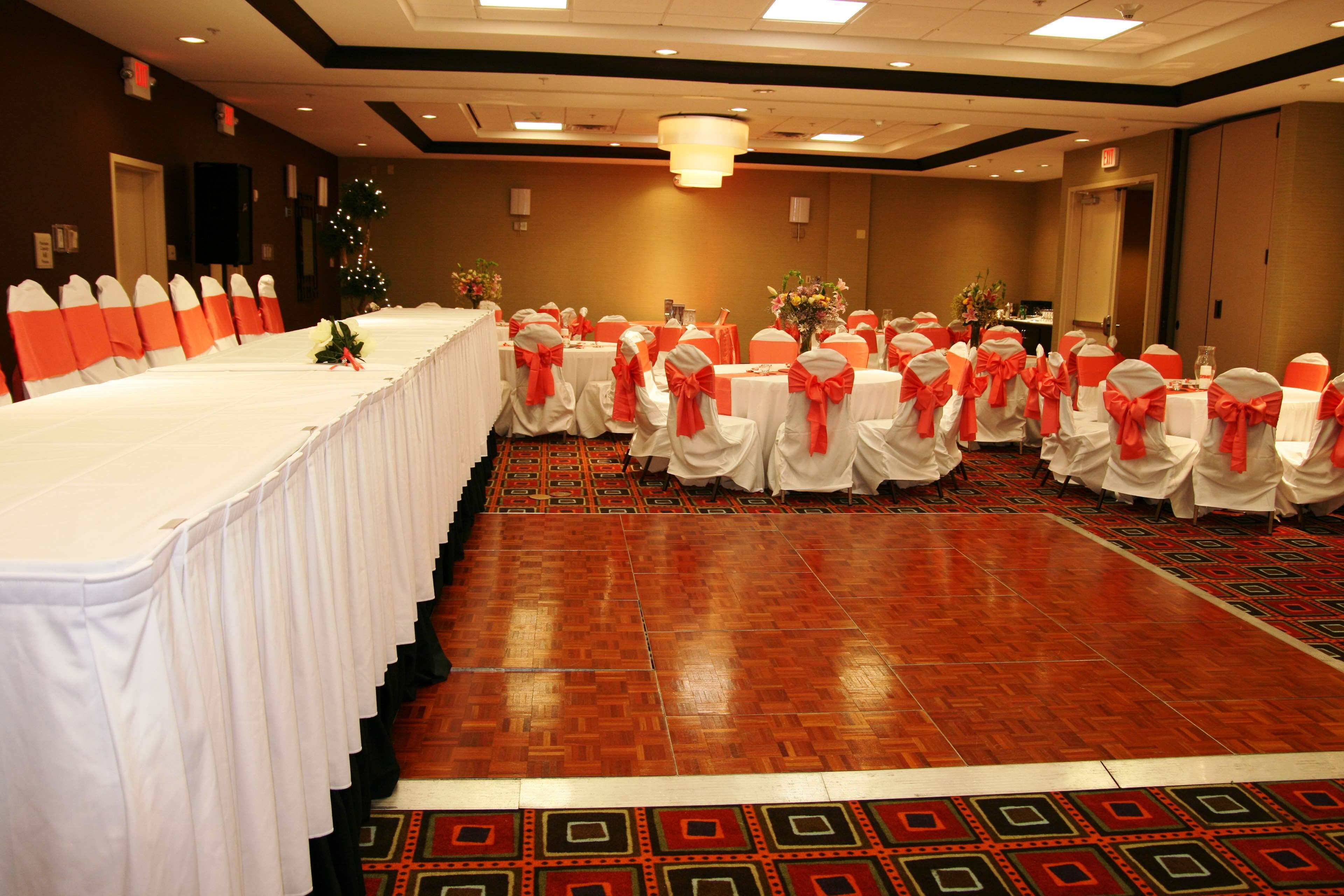 Un salón de banquetes con filas de mesas cubiertas con manteles blancos y sillas adornadas con cintas naranjas.
