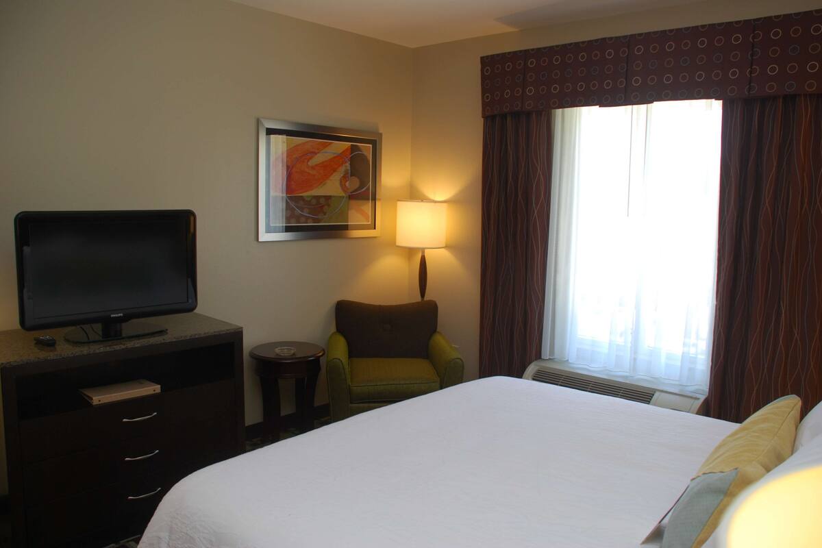 Habitación de hotel con cama, televisión sobre una cómoda, una silla, una lámpara y ventana con cortinas.