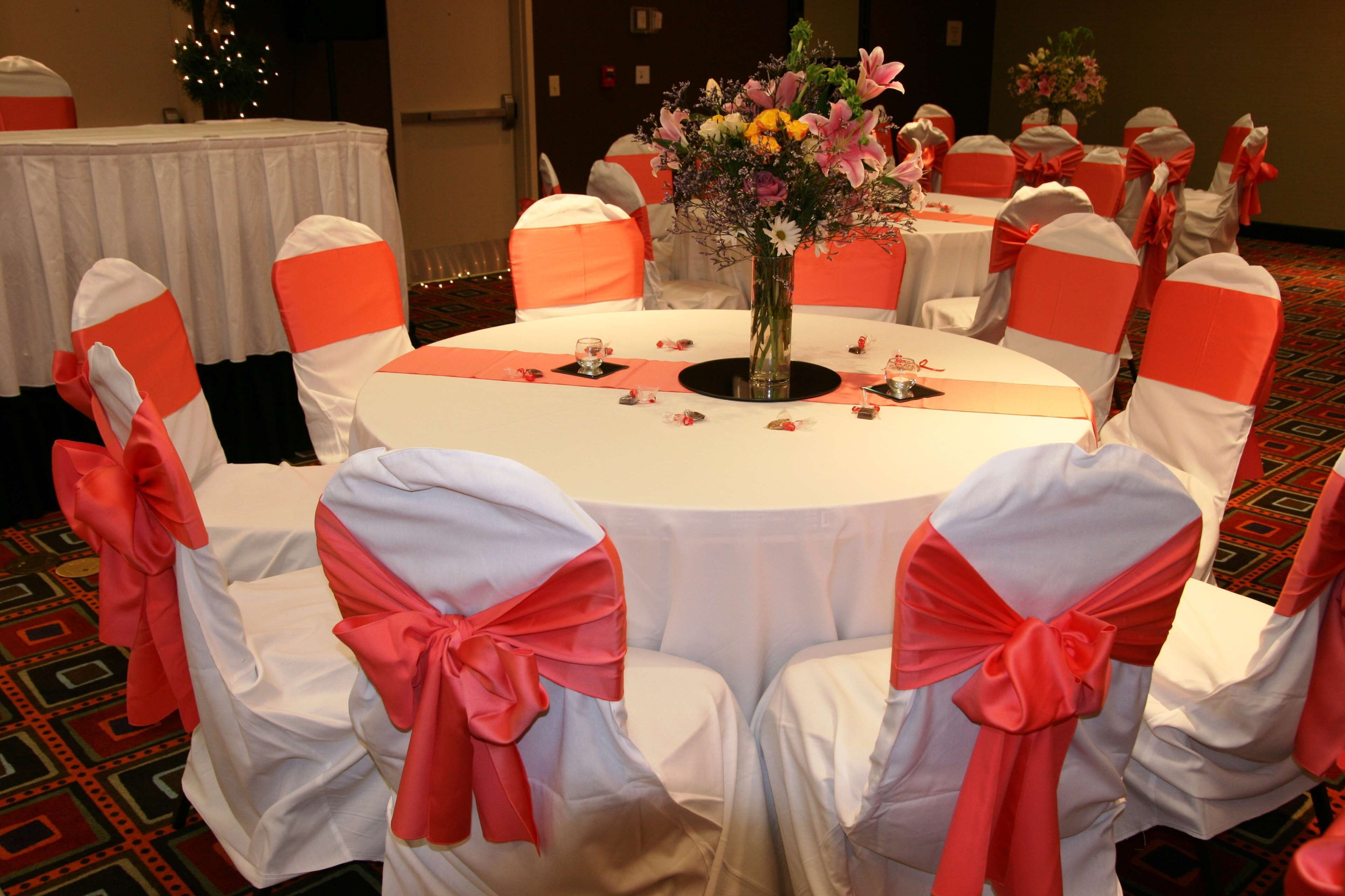 Une table ronde recouverte d’une nappe blanche, entourée de chaises habillées de housses blanches et de rubans rouges, et un vase rempli de fleurs.