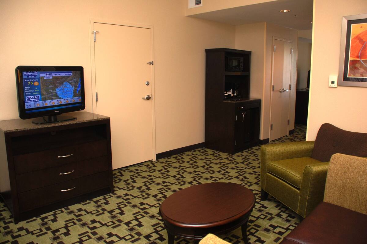 Habitación de hotel con una televisión de pantalla plana mostrando un pronóstico del tiempo, un aparador de madera, un sillón verde, un sofá marrón y una alfombra estampada.