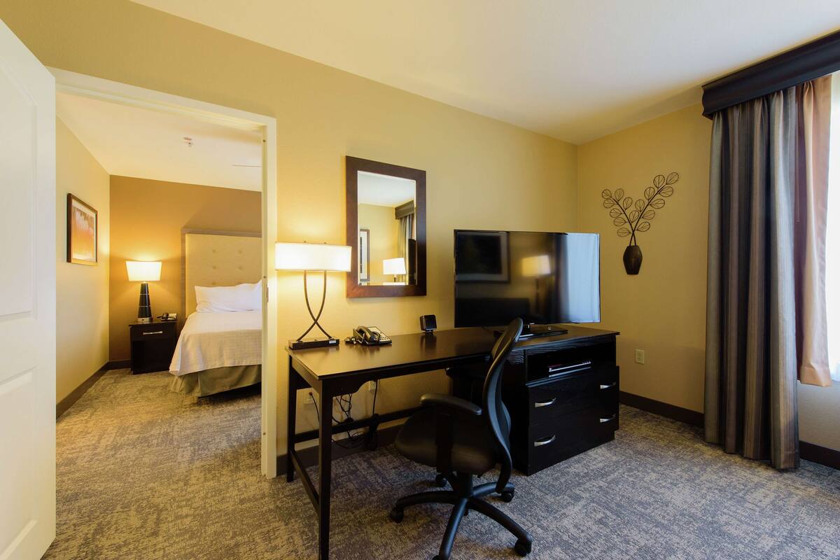 A hotel room with a bed, desk, chair, TV, and a small nightstand with a lamp.