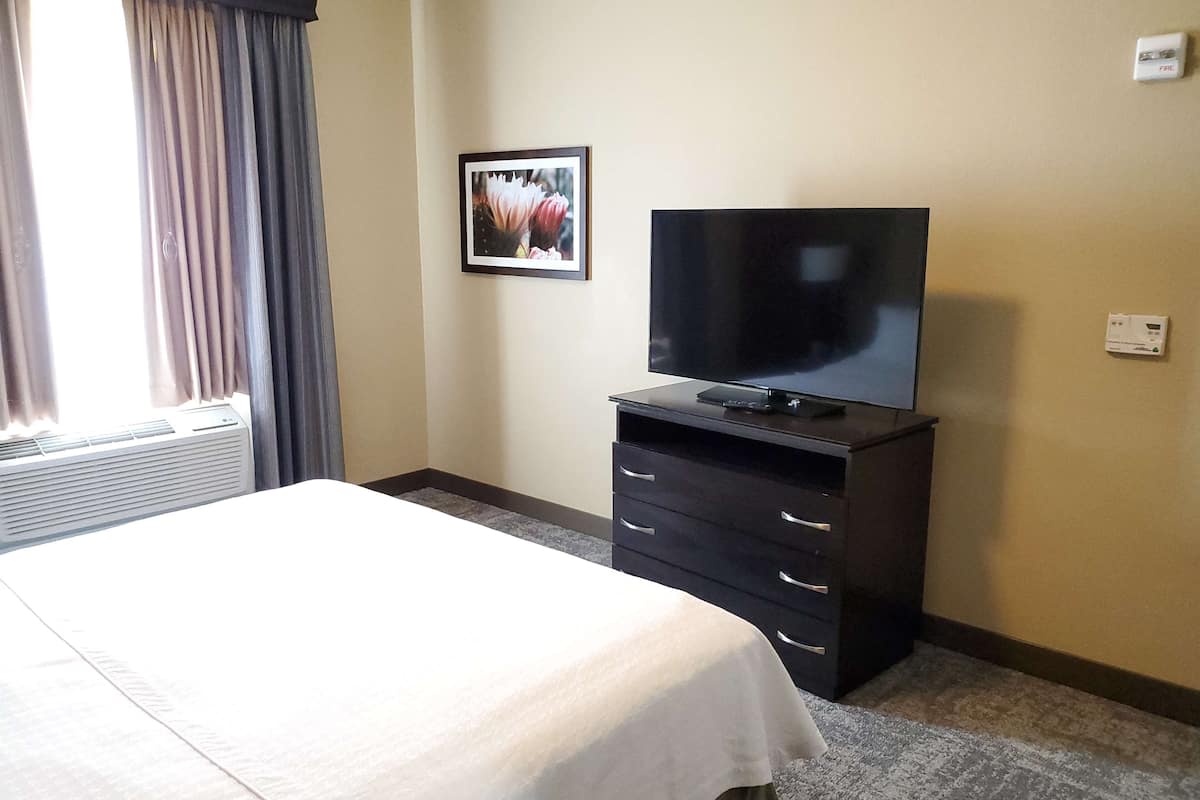 A hotel room with a bed, a TV on a dresser, a framed picture on the wall, and a window with curtains.
