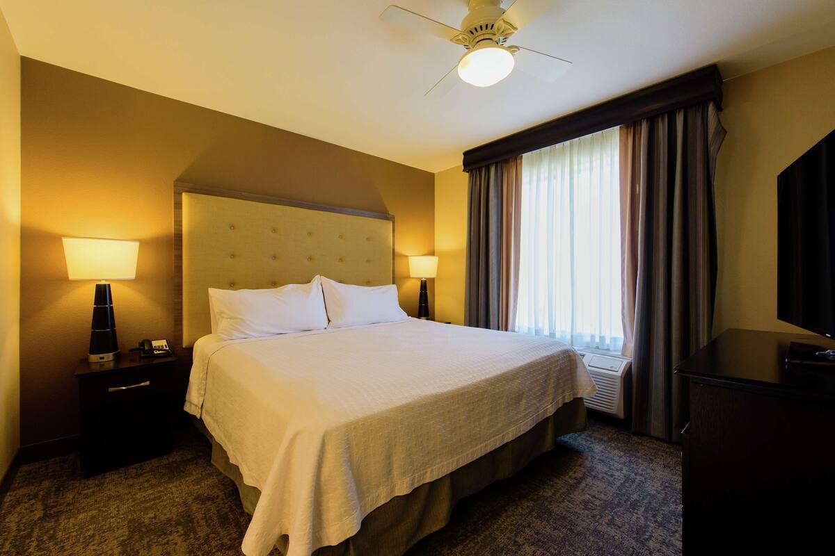 A hotel room with a large bed, two bedside tables with lamps, a ceiling fan, a flat-screen TV, and a window with curtains.