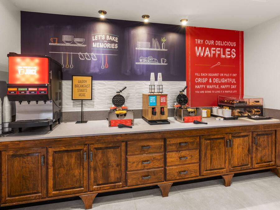 A breakfast bar with waffle makers, a juice machine, and a sign promoting waffles.